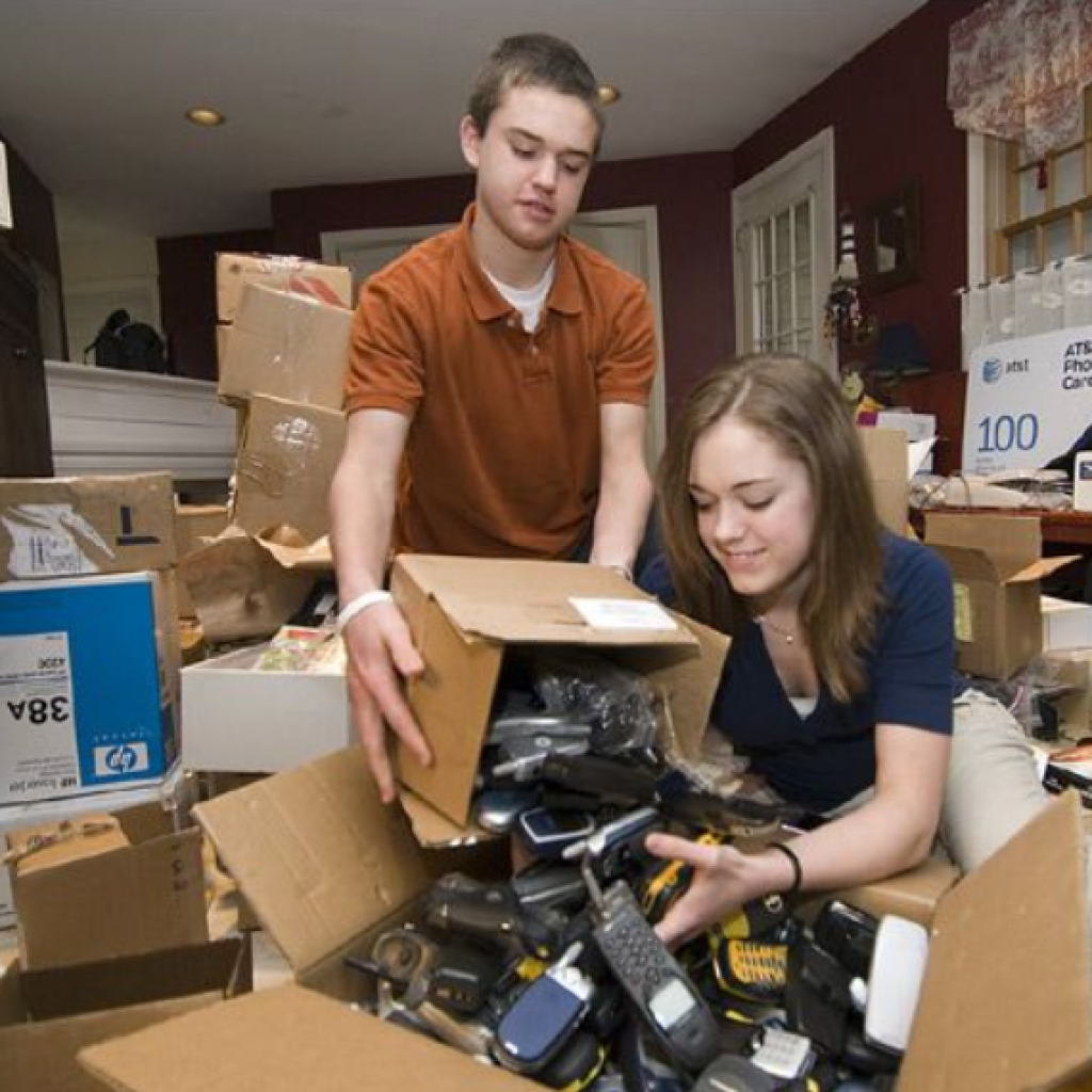 Two young volunteers sorting a large box of donated old cell phones for recycling